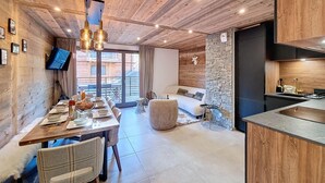 A modern interior with a wooden ceiling, a dining area with a table set for four, a stone accent wall, and a kitchen with a sink and stove. - Le Grand Cerf - appartement 8 pers - proche centre (Les Gets)