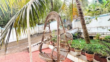Una silla colgante de mimbre con estructura circular, ubicada en un patio de ladrillo rojo con plantas en macetas y una palmera al fondo.