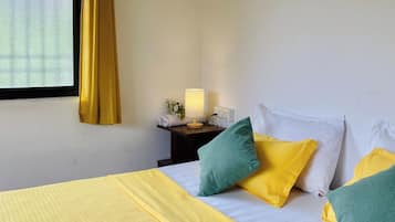 Una habitación de hotel con cama, almohadas, mesita de noche, lámpara y ventana con cortinas.