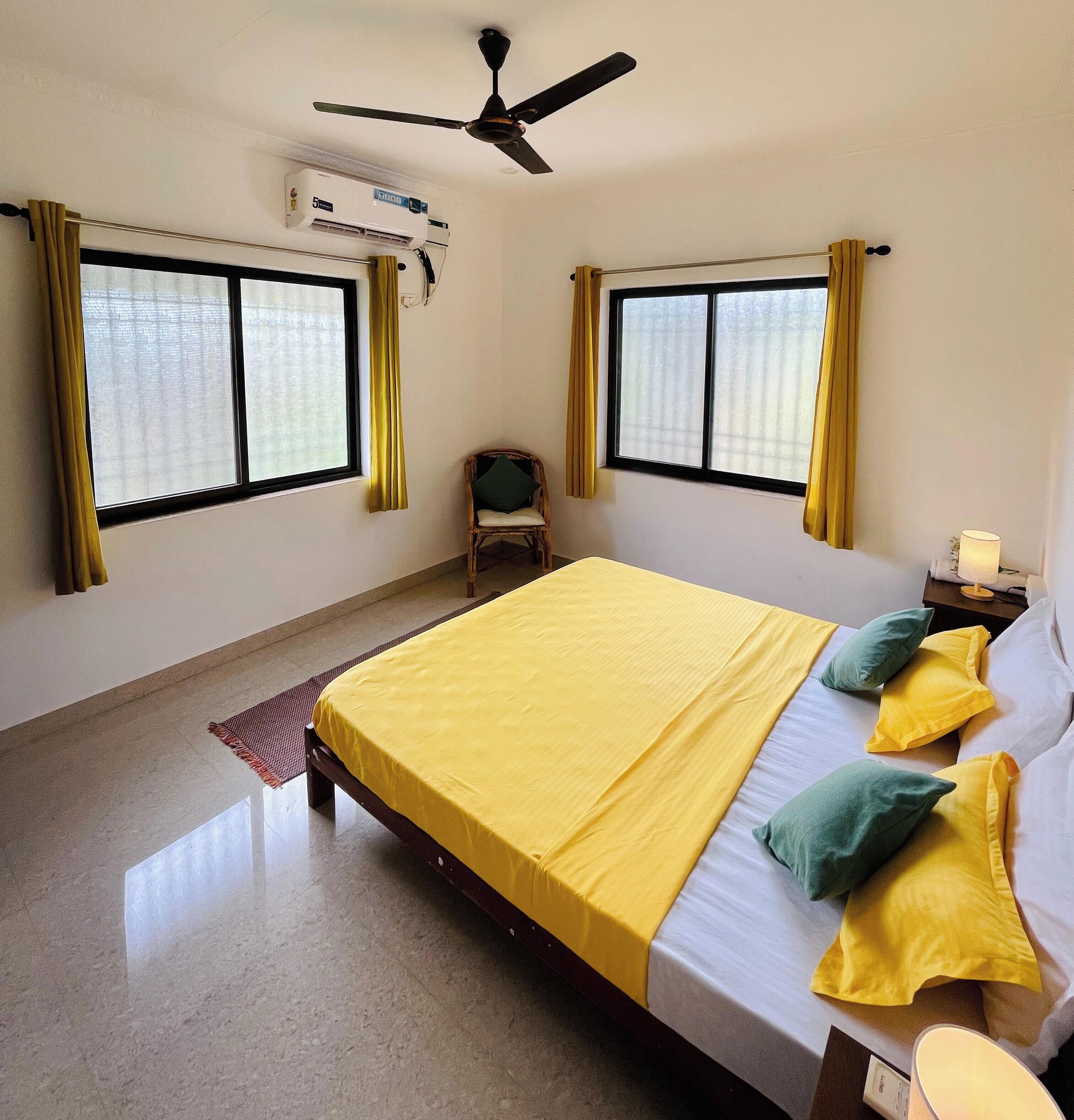 A bedroom with a bed, two windows, an air conditioner, a ceiling fan, and a chair.