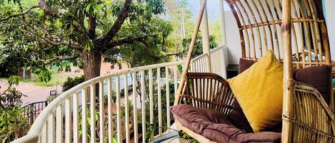 A wicker swing chair with cushions on a balcony with a white railing, overlooking a garden with trees and a clear sky.