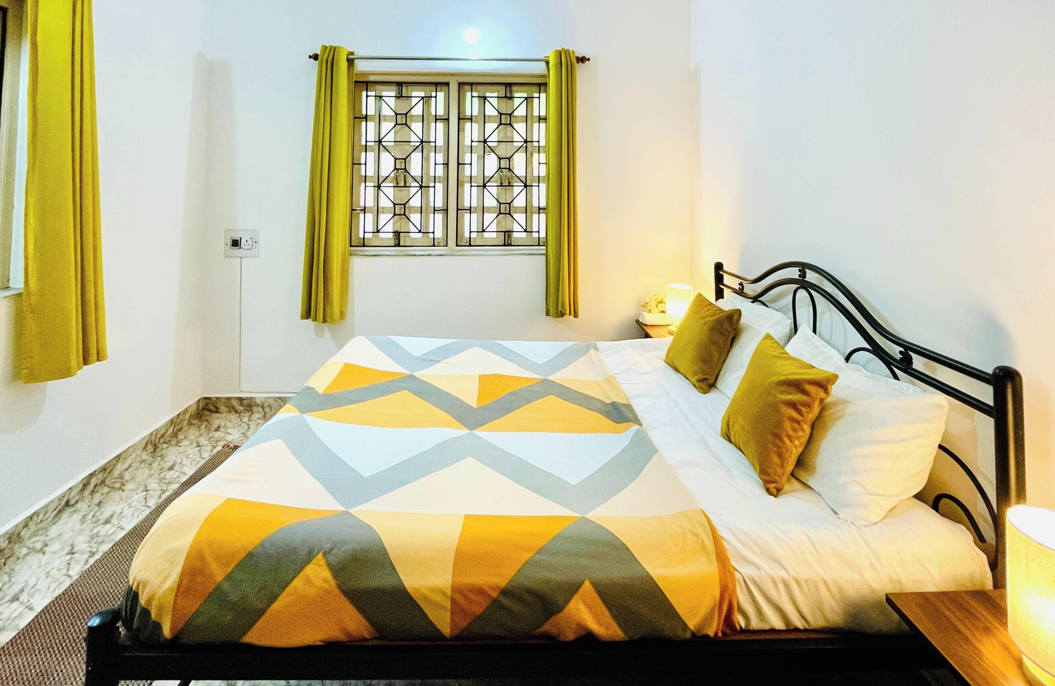 A bed with a geometric patterned bedspread, a bedside table with a lamp, and a window with a metal grille and green curtains.