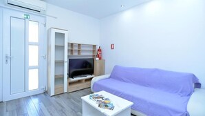 A room with a purple sofa, a white coffee table with magazines, a TV on a wooden stand, and a white door with a green exit sign. - Lovely flat just steps from the beach (Fažana)
