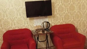 Une pièce avec un papier peint à motifs, un téléviseur à écran plat fixé au mur, une petite table avec une bouilloire et une tasse, et deux fauteuils rouges.