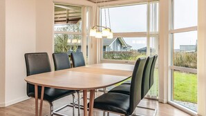Un espace repas avec une table en bois et des chaises noires, de grandes fenêtres donnant sur des maisons et de la verdure, et un luminaire suspendu.