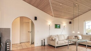 Un salon spacieux avec un plafond en bois, un canapé blanc, une table basse en verre et un tableau accroché au mur.