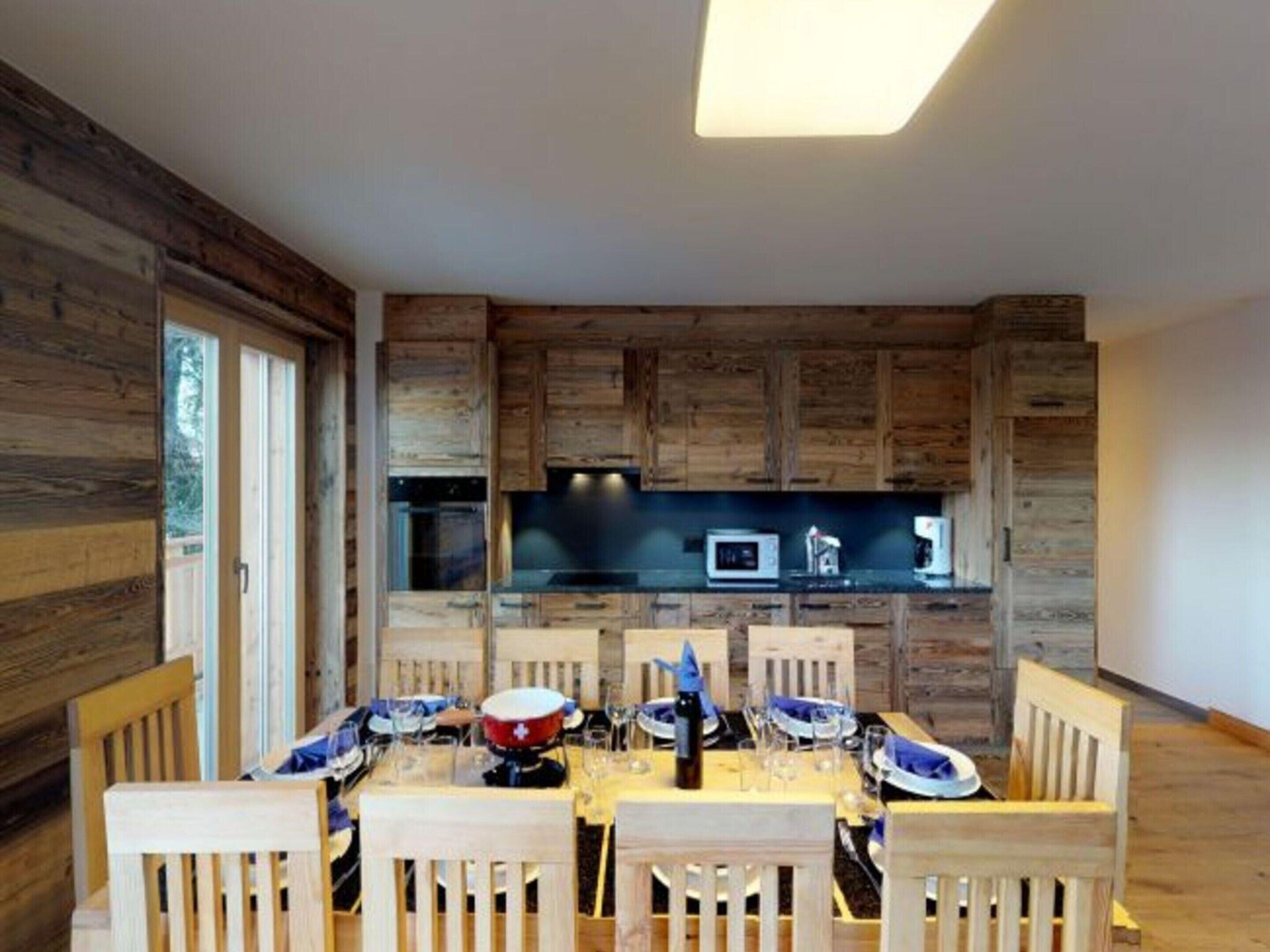 A dining area with a wooden table set for four, surrounded by wooden chairs. A kitchen with wooden cabinets and a microwave is visible in the background.