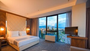 Ein Hotelzimmer mit einem großen Bett, einem Fernseher und Blick auf eine Stadtlandschaft durch große Fenster.