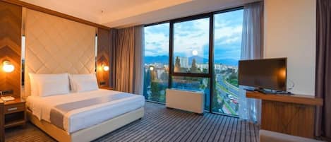 A hotel room with a large bed, a TV, and a view of a cityscape through large windows.
