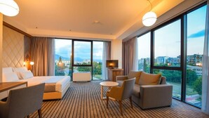 Ein Hotelzimmer mit Bett, Sofa, kleinem Tisch, Fernseher und großen Fenstern mit Blick auf die Stadt.