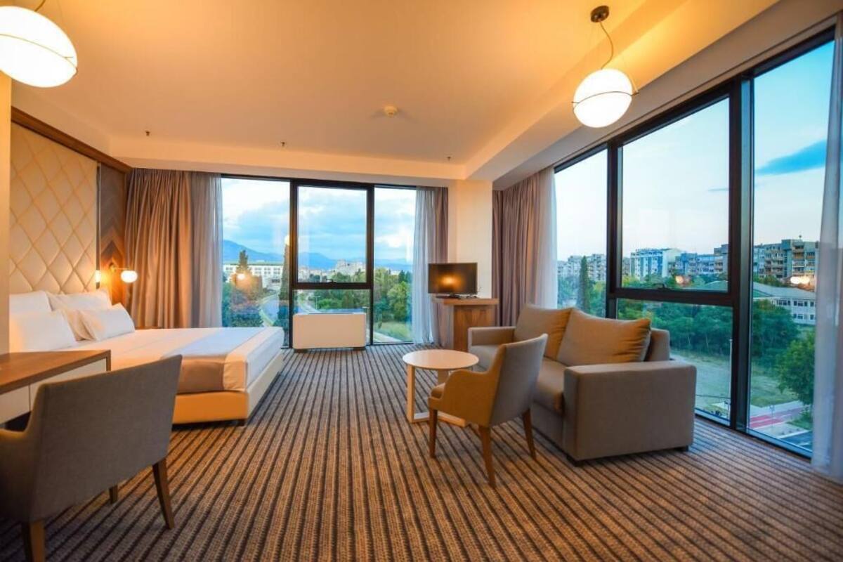 A hotel room with a bed, a sofa, a small table, a TV, and large windows offering a city view.