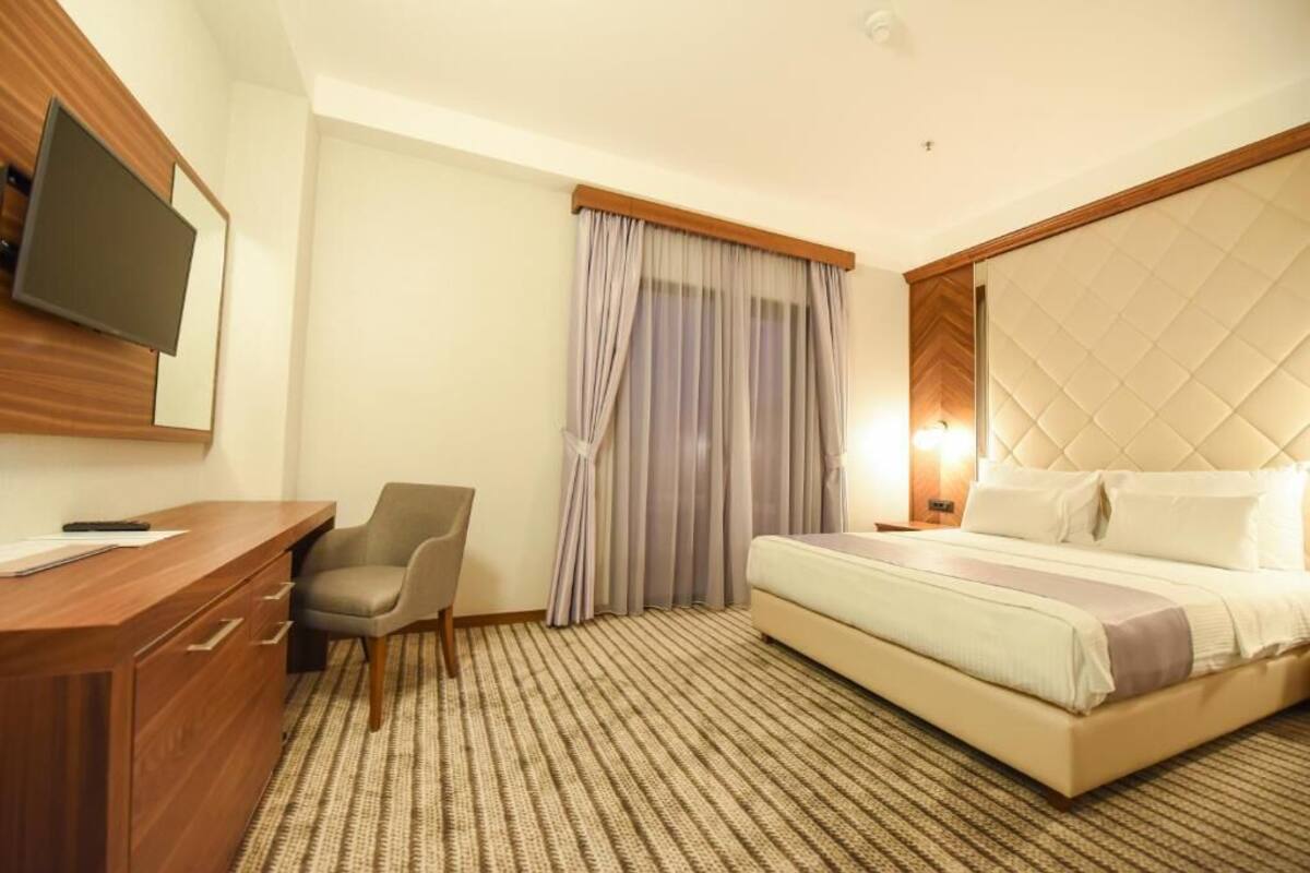 A hotel room with a bed, a desk with drawers, a chair, a television mounted on the wall, and a window with curtains.