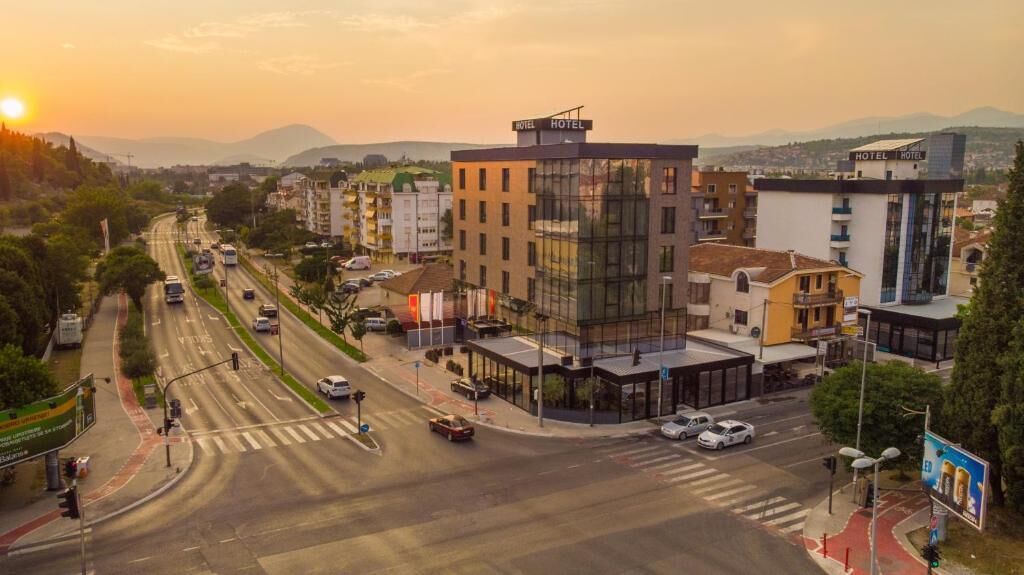 A modern hotel with a glass facade, located at a city intersection with visible traffic and surrounding buildings.