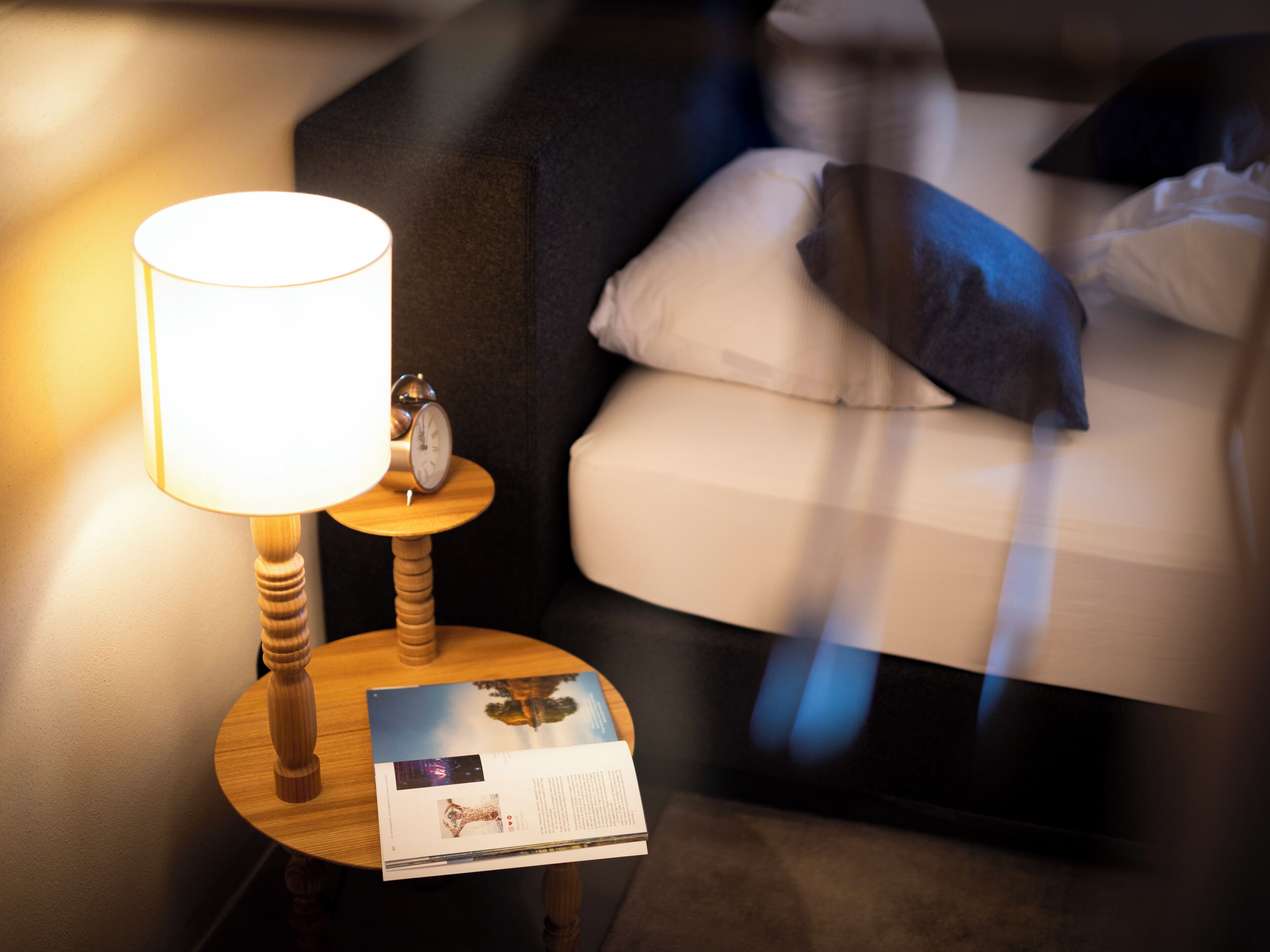 A neatly made bed with pillows, a bedside lamp, and a small table with a clock and a brochure.