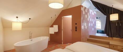 A modern bedroom with a large white bathtub, a bed with white linens, and a staircase leading to an upper level.