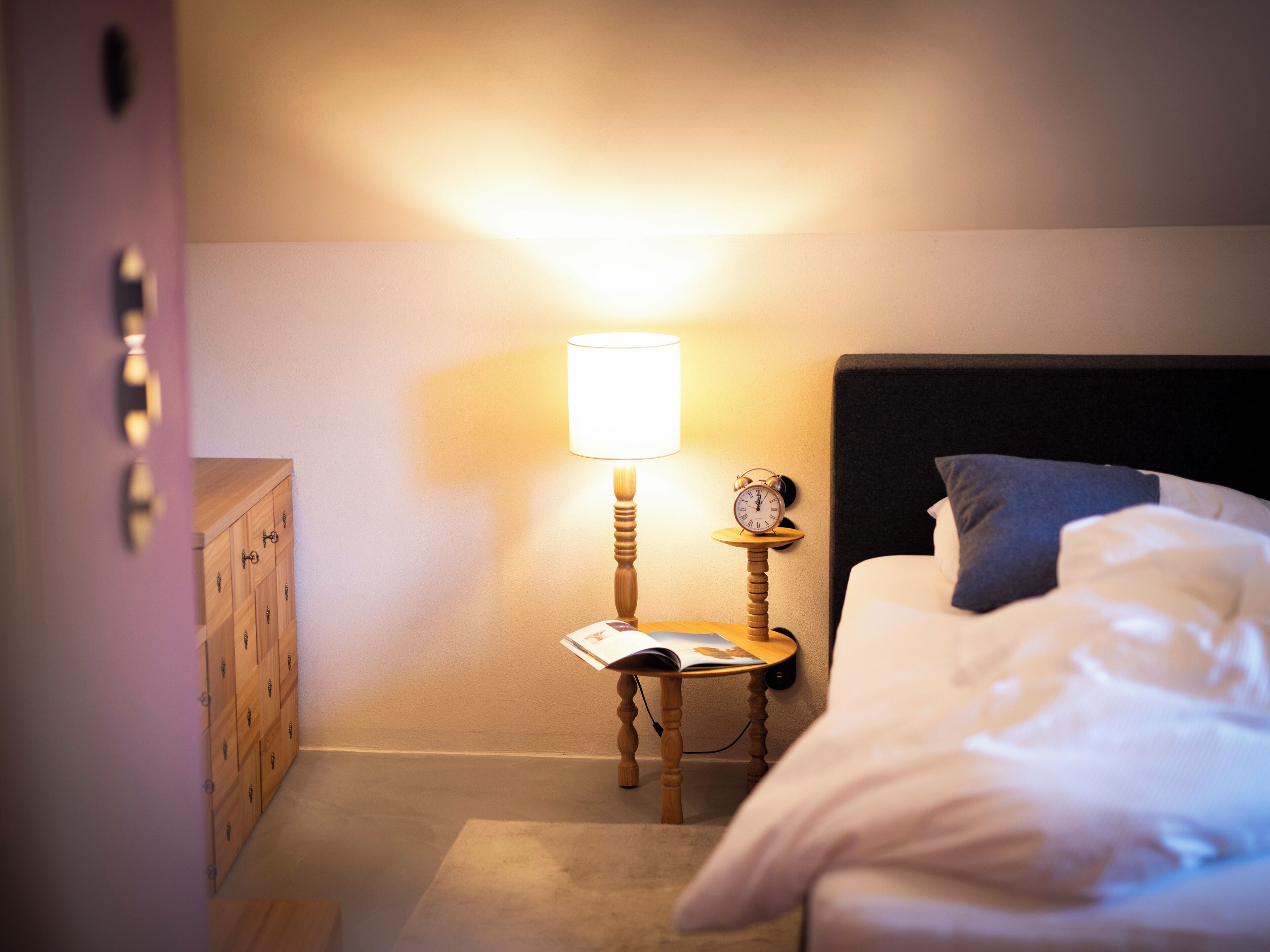 Une chambre d’hôtel avec un lit, une table de chevet avec une lampe, une horloge et une commode en bois.