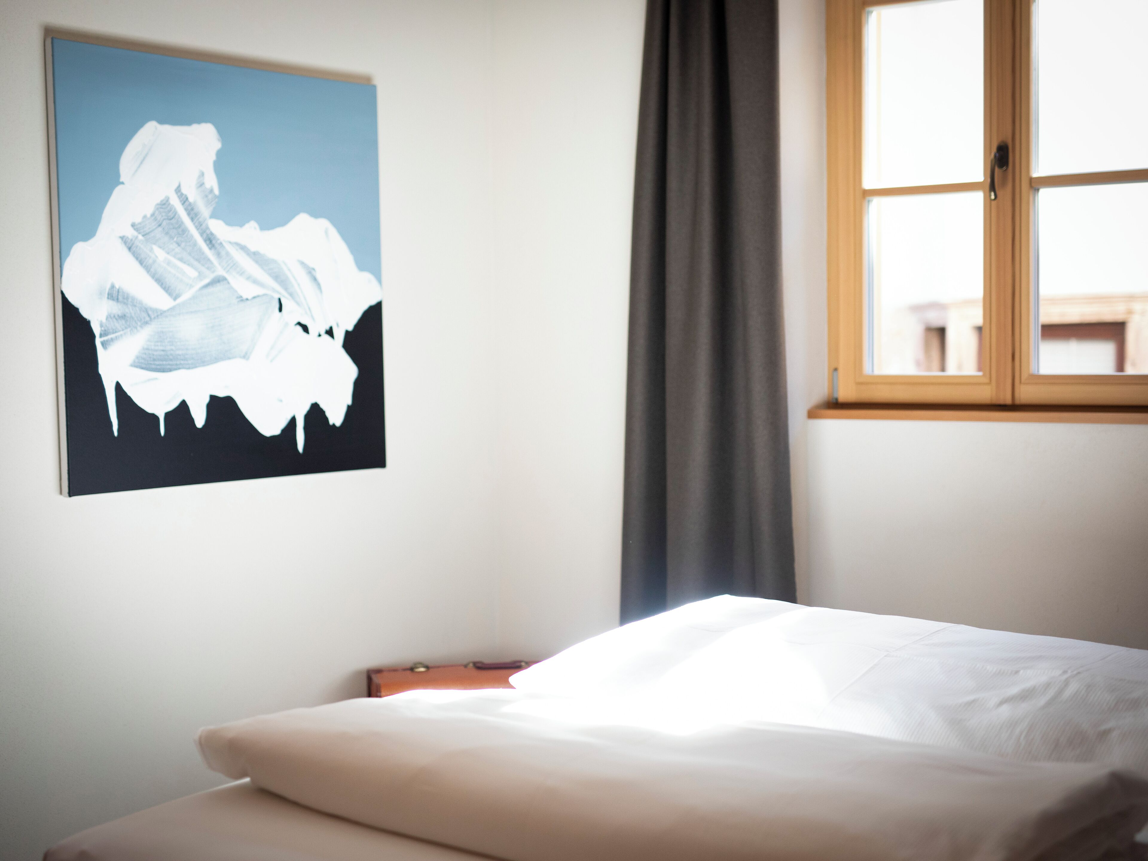 A hotel room with a bed, a window with curtains, and a wall-mounted painting.