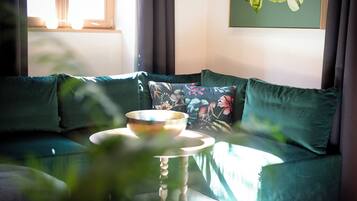 Un canapé vert agrémenté de coussins à motifs floraux, un bol posé sur une table et un tableau accroché au mur.