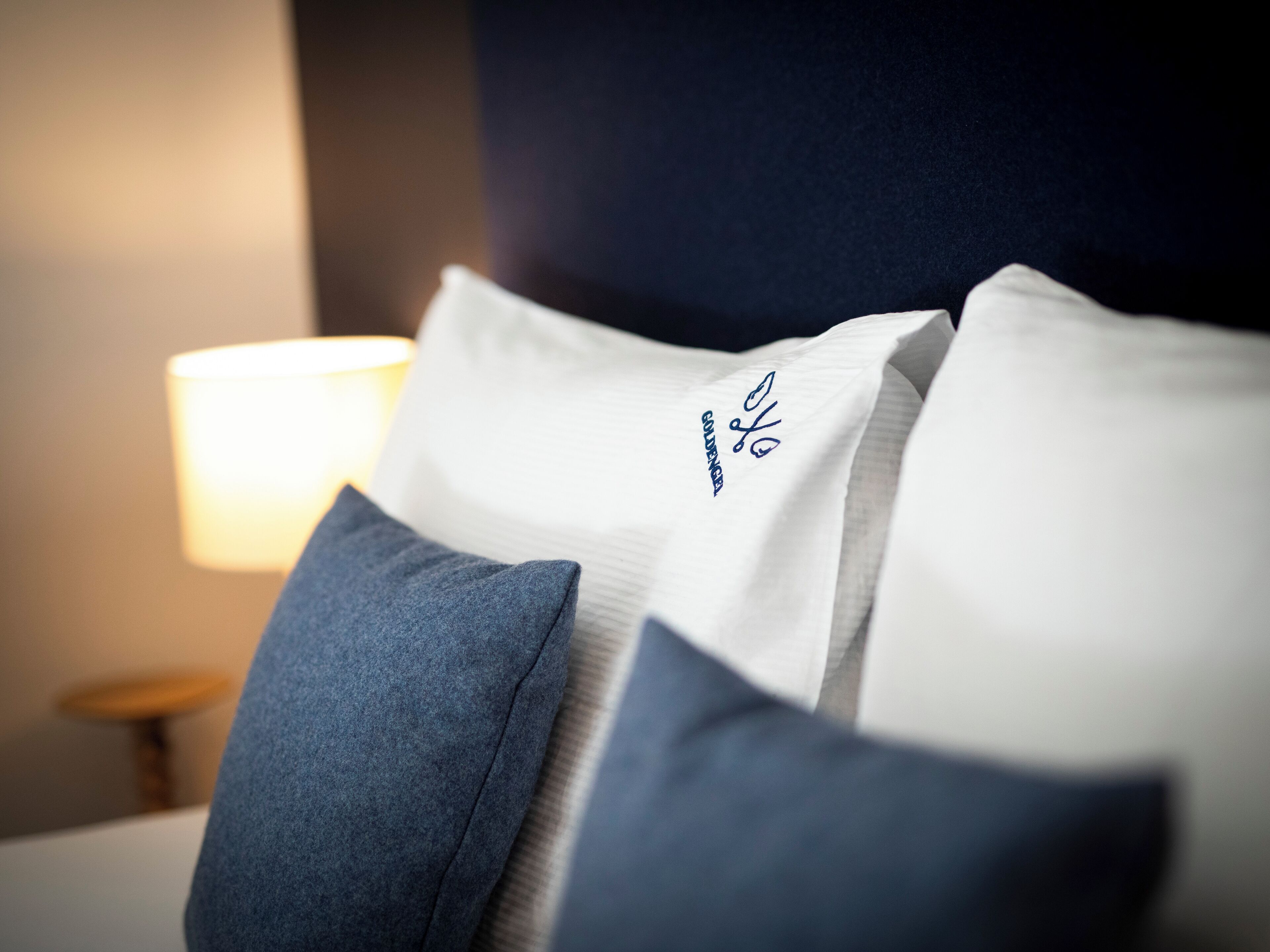A close-up of a bed with white and blue pillows, a lamp in the background, and a dark wall.