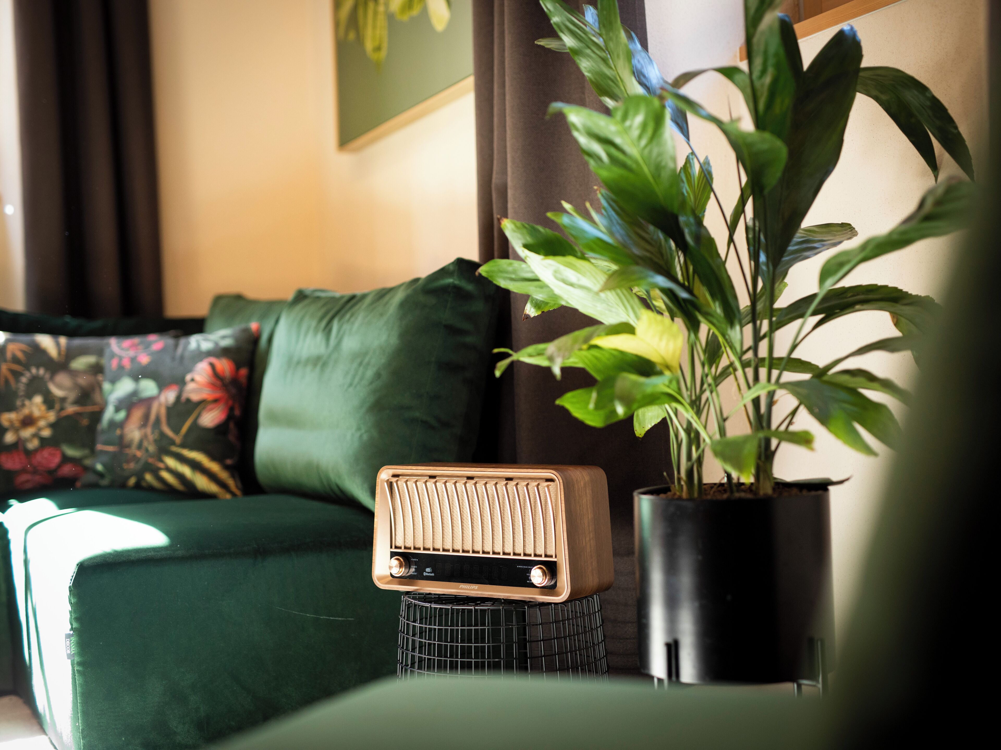 Ein grünes Sofa mit floralen Kissen, ein kleiner Tisch mit einem Vintage-Radio und ein Topfpflanzen.