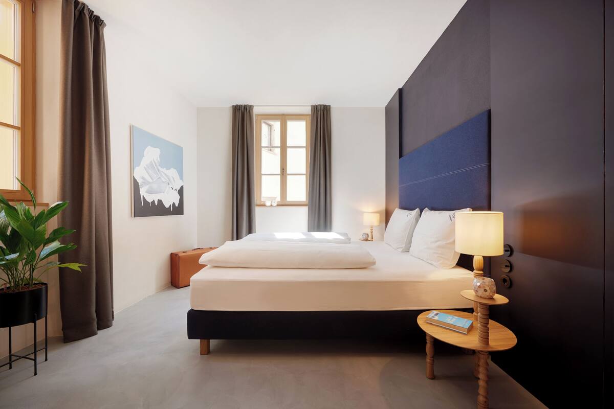 A hotel room with a bed, bedside table, lamp, and a potted plant.