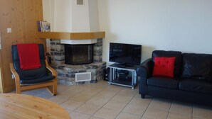 Un salon doté d’une cheminée en pierre, d’un canapé en cuir noir agrémenté de coussins rouges, d’une chaise en bois avec un plaid rouge, et d’un téléviseur posé sur un meuble.
