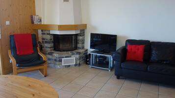 A living room with a stone fireplace, a black leather sofa with red cushions, a wooden chair with a red throw, and a television on a stand.