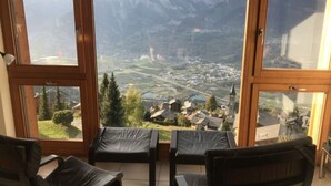 A room with a large window offering a view of a town and mountains. There are two black leather chairs and a small table. - Mountain Apartment With Parking (Veysonnaz)