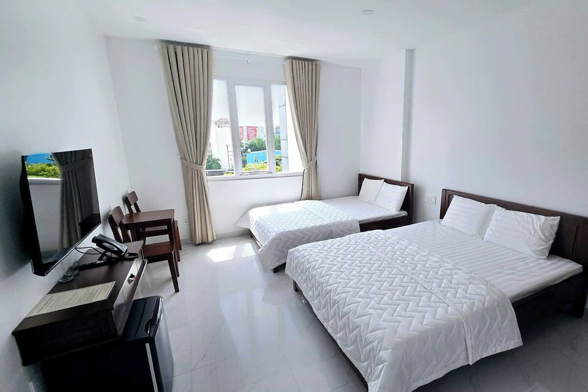 A hotel room with two beds, a desk, a chair, a TV, and large windows with curtains.