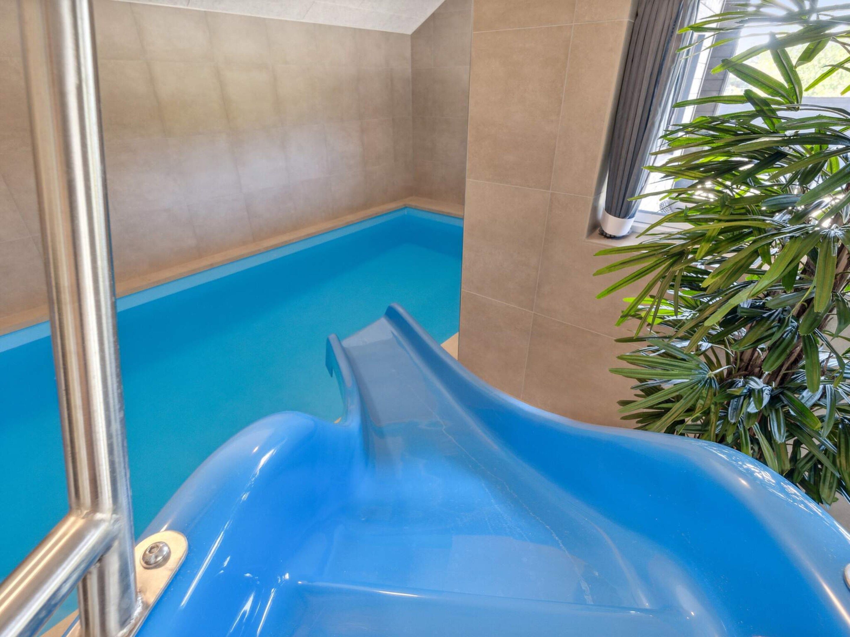 A blue indoor water slide with a metal handrail, surrounded by beige tiled walls and a window with natural light.