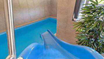 A blue indoor water slide with a metal handrail, surrounded by beige tiled walls and a window with natural light.