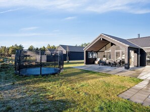Un patio exterior moderno con un trampolín, un área de estar cubierta y un edificio con ventanas amplias.