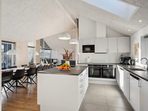 A modern kitchen with white cabinetry, a central island with fruit, and a dining area with chairs and tables.