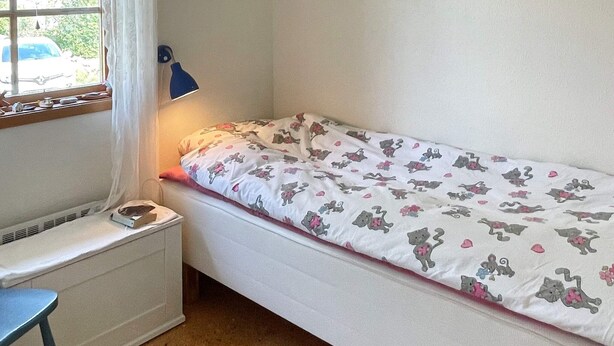 Quarto pequeno com cama de solteiro, janela, escrivaninha e luminária.