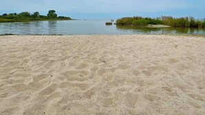 Uma praia arenosa com águas calmas, um litoral distante e alguma vegetação.