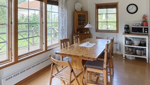 Mesa de jantar de madeira com cadeiras, um relógio e eletrodomésticos na cozinha, num espaço com grandes janelas.