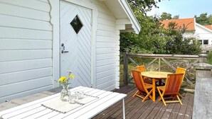 A wooden deck with a white table and orange chairs, a white door with a diamond-shaped window, and a wooden railing. - 8 Person Holiday Home in Borgholm (Borgholm)