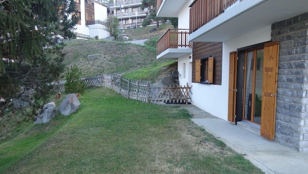 Une maison moderne à deux étages, dotée d’une façade en pierre, de balcons en bois et d’un petit jardin clos par une clôture en bois, avec un arbre.