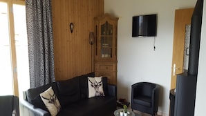 Un salon avec un canapé en cuir noir, un meuble en bois, un fauteuil noir, une petite table avec une plante et un téléviseur noir fixé au mur.