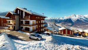 A multi-story hotel with balconies, a car parked in front, and snow-covered surroundings with mountains in the background. - Stunning Apartment With Valley and Mountains (Veysonnaz)