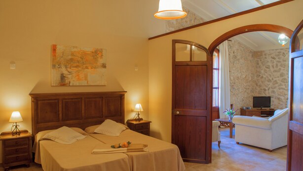 A hotel room with a large bed, two bedside tables with lamps, a wooden door, and a view of a living area with a sofa and a TV. - Agroturismo Roqueta (Maria de la Salut)
