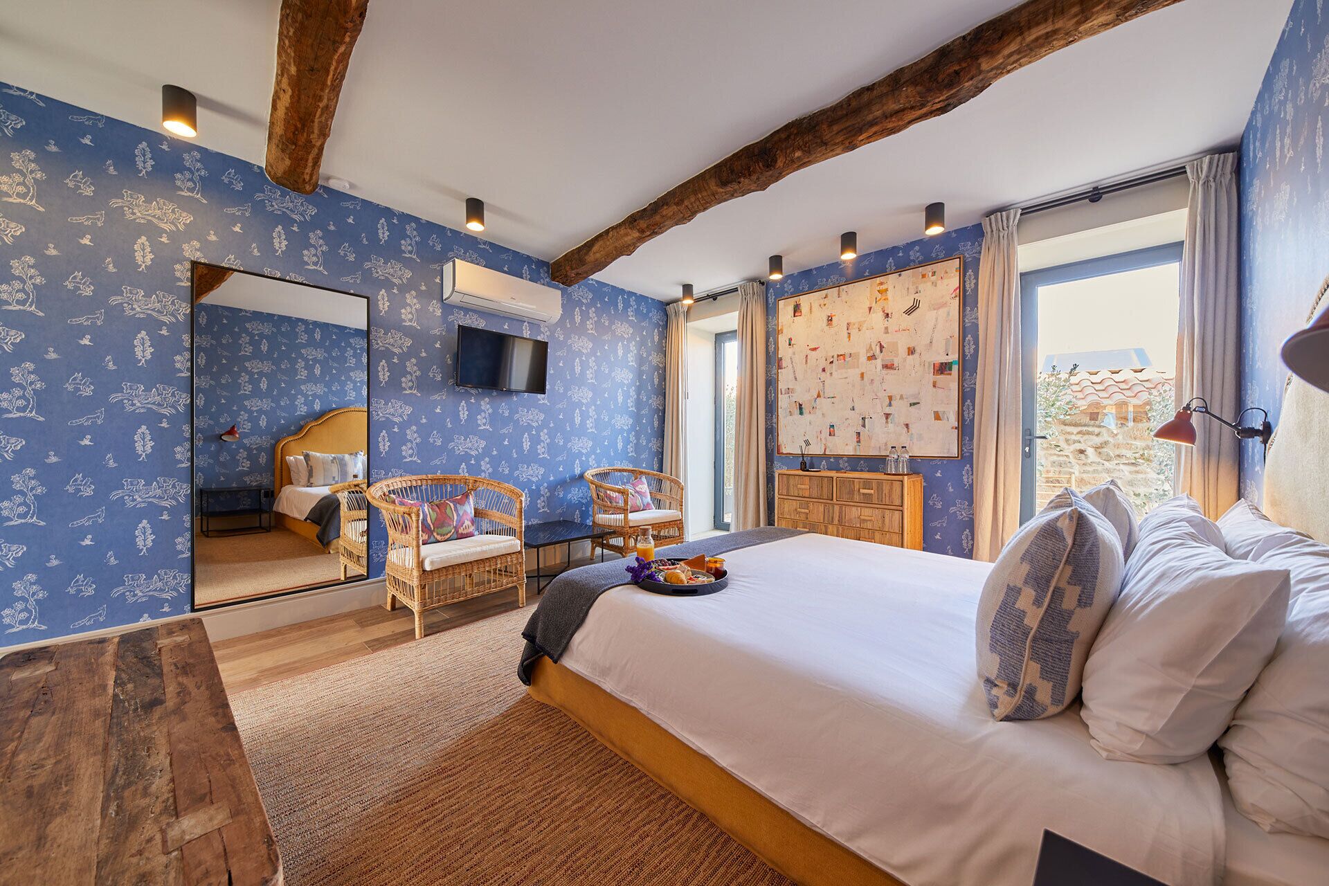 A bedroom with a large bed, two wicker chairs, a wooden cabinet, a flat-screen TV, and a large window with curtains.
