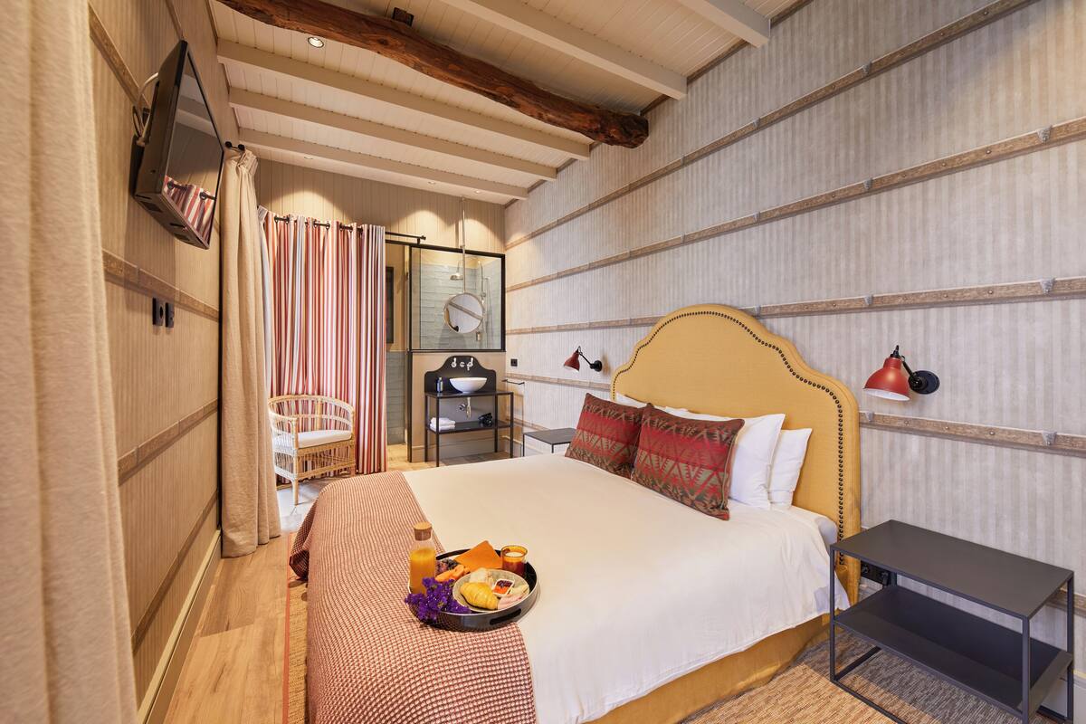Ein Hotelzimmer mit einem großen Bett, einem Nachttisch, einem kleinen Tisch, einem Sessel, einem Spiegel, einem Regal und einem Fenster.