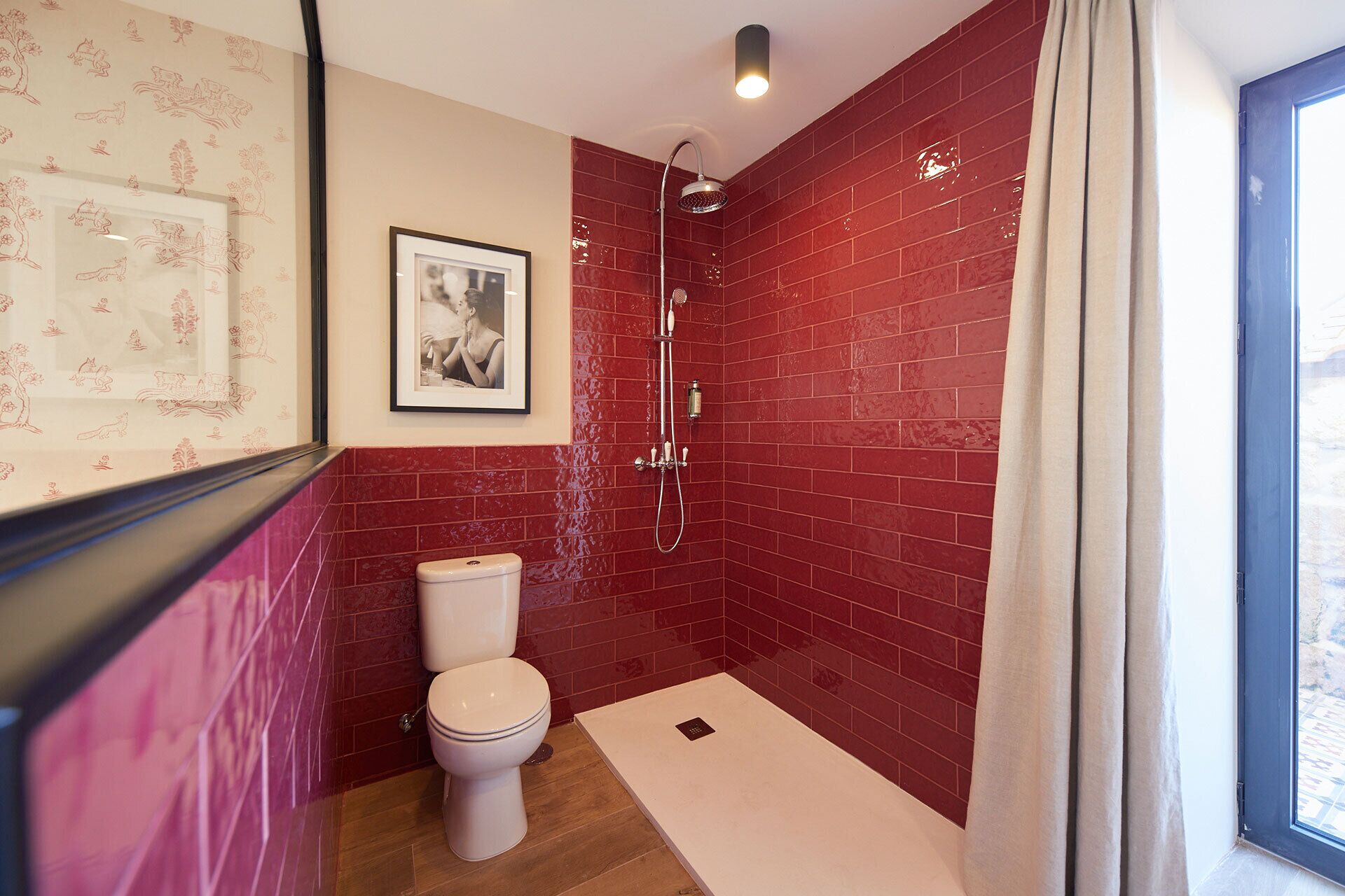 Un baño con paredes de azulejos rojos, un inodoro blanco, un cuadro en la pared y una ventana con cortina.
