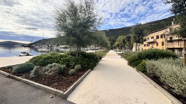 Eine Küstenpromenade mit freiem Blick auf das Meer, ein Hafen mit Booten und Gebäude mit Balkonen zum Wasser hin.