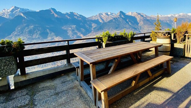 Ein Holztisch mit Bänken, umgeben von einer Steinmauer und einem Holzgeländer, mit Blick auf ein Gebirge.