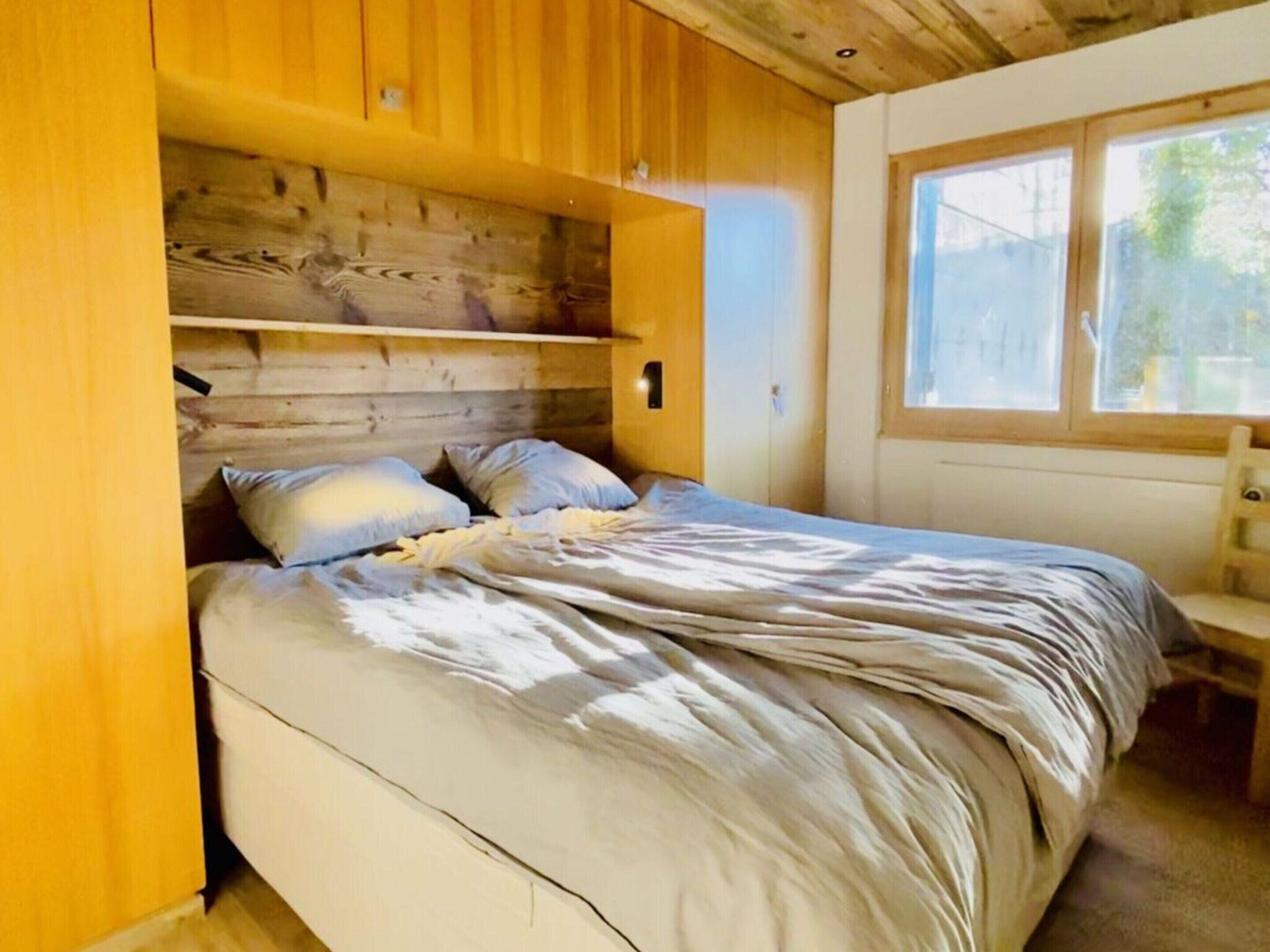 A bedroom with a wooden headboard, a bed with a grey duvet, and a window providing natural light.