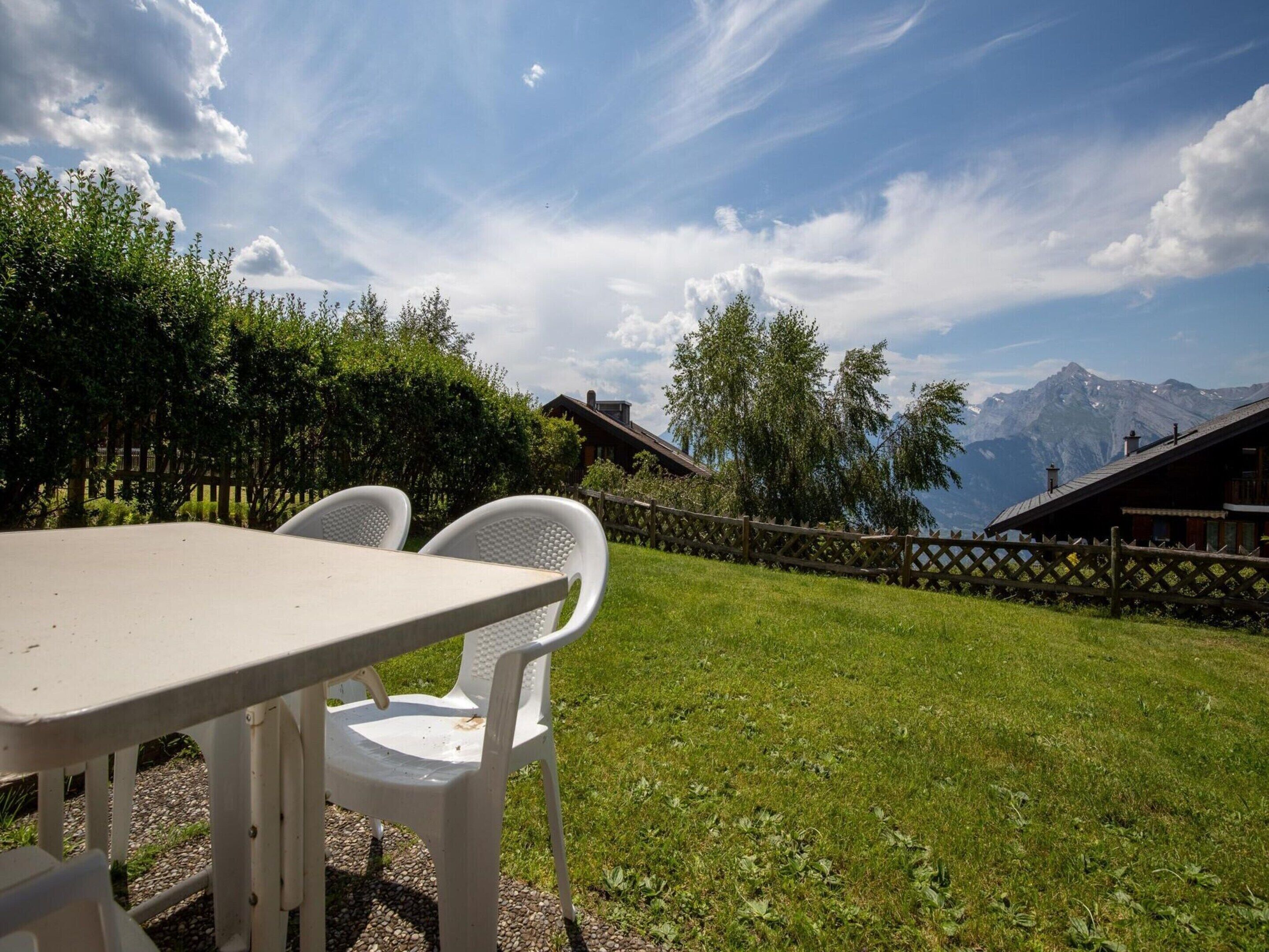 A table with two white chairs in a grassy outdoor area with a mountain view.