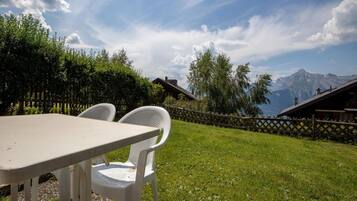 A table with two white chairs in a grassy outdoor area with a mountain view.