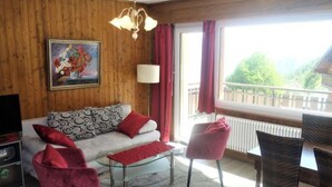 A cozy living room with a wooden wall, a sofa with patterned cushions, a glass coffee table, red chairs, and a painting on the wall. - Beaulieu F 014 Center Apartment 5 Pers (Veysonnaz)